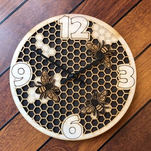 May include: A round wooden clock with a honeycomb design and three decorative bees. The clock face features large, cutout numbers at the 12, 3, 6, and 9 positions. The clock hands are black.