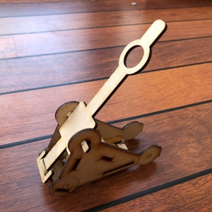 Op de afbeelding: Een klein, onafgewerkt houten katapultspeelgoed met een hendel en wielen. De katapult is gemaakt van lichtgekleurd hout en heeft een cirkelvormige opening aan het einde van de hendel. Het speelgoed is ontworpen om kleine voorwerpen te lanceren.