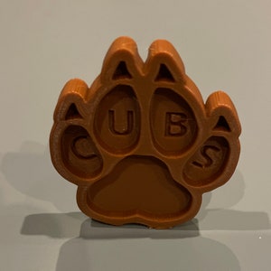 May include: A brown, 3D-printed paw print with the letters "CUBS" embedded within the pads. The paw print is a solid, textured object, likely made of plastic or a similar material. The design is a stylized representation of a bear's paw.