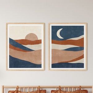 Puede incluir: Dos impresiones abstractas de paisajes enmarcadas con un motivo de sol y luna. Las impresiones están colgadas sobre una cama con una funda nórdica marrón y almohadas a rayas. La cama está en un dormitorio con mesitas de noche de madera y lámparas colgantes.