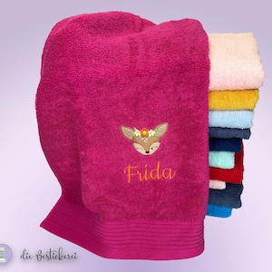 Könnte beinhalten: Ein fuchsiafarbenes Badetuch mit einem gestickten Hirschgesicht mit Blumenkranz und dem Namen "Frida" in Goldfaden. Rechts ist ein Stapel bunter Handtücher zu sehen.