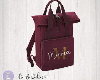 Könnte beinhalten: Weinroter Rucksack mit Rollverschluss und verstellbaren Trägern. Der Rucksack hat einen goldfarbenen Schriftzug "Mama" auf der Vorderseite.
