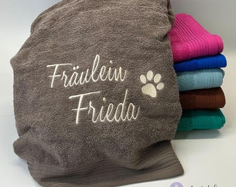 May include: A brown towel with the embroidered text "Fräulein Frieda" and a paw print. The towel is folded and stacked on top of other towels in various colors.
