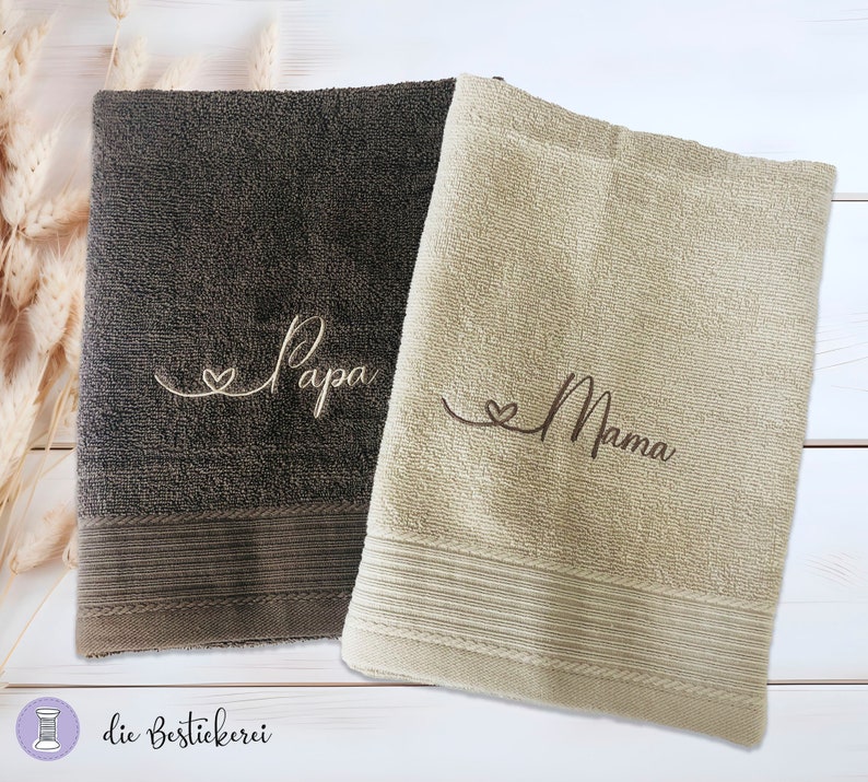 May include: Two embroidered towels, one brown and one beige, with the words "Papa" and "Mama" in cursive script. Each towel has a small heart symbol before the word.