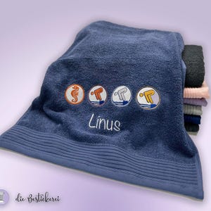 Könnte beinhalten: Marineblaues Badetuch mit gestickten Designs zum Thema Schwimmen und dem Namen "Linus" in Weiß. Das Handtuch zeigt ein Seepferdchen und Schwimmer in Kreisen. Ein Stapel gefalteter Handtücher ist im Hintergrund zu sehen.