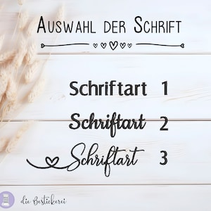 May include: A white wooden background with three different font styles displayed. The text reads "Auswahl der Schrift" with hearts and a line underneath. The text below reads "Schriftart 1", "Schriftart 2", and "Schriftart 3".