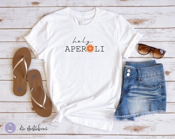 May include: White t-shirt featuring the text "holy APEROLi" and an orange slice graphic. The shirt is styled with denim shorts, sunglasses, and sandals.