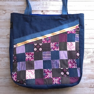 Può includere: Una borsa tote in denim blu con un design a patchwork con tessuti viola, rosa e marrone. La borsa ha una cerniera dorata e una tracolla in denim blu.