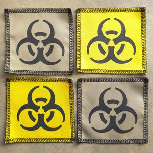 Può includere: Quattro toppe quadrate con un simbolo di rischio biologico nero su sfondo giallo o beige. Le toppe hanno un bordo nero cucito.