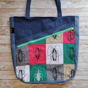 Könnte beinhalten: Eine blaue Jeans-Tragetasche mit einer Reißverschlusstasche und einem bunten Patchwork-Design mit Schwarzweiß-Illustrationen von Kakerlaken. Die Tasche hat einen grünen Reißverschluss und einen goldenen Reißverschluss-Zipper.