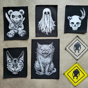 Könnte beinhalten: Eine Sammlung von schwarz-weißen Aufnähern mit gruseligen Designs. Dazu gehören ein Teddybär mit Messer, ein Geist, ein Schädel mit Hörnern, eine Katze, ein katzenartiges Wesen und zwei Warnschild-Aufnäher mit einer Monstersilhouette.