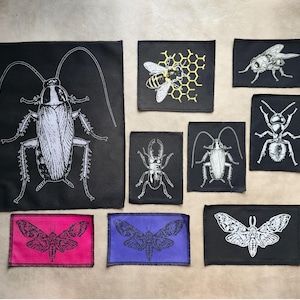 Op de afbeelding: Een verzameling zwarte stoffen patches met witte insectenillustraties. De patches tonen een kakkerlak, een bij, een vlieg, een mier, een kever en motten. Sommige patches hebben roze of paarse randen.