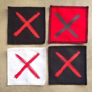 Può includere: Quattro toppe di tessuto con una X rossa su sfondo nero o bianco. Le toppe sono quadrate e hanno un bordo cucito.