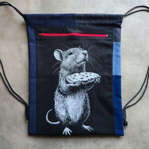 Può includere: Zaino con coulisse con un pannello frontale nero con un'illustrazione dettagliata di un ratto che tiene in mano un pezzo di formaggio. La borsa ha una cerniera rossa, dettagli in denim e coulisse nere.