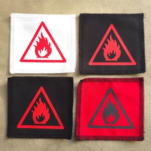 Op de afbeelding: Vier stoffen patches met een rode vlam in een zwarte driehoekige waarschuwingsbord. De patches zijn wit, zwart, zwart met witte stiksels en rood met witte stiksels.