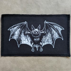 Può includere: Toppa rettangolare nera con un'illustrazione bianca dettagliata di un pipistrello con le ali spiegate. Il pipistrello ha un volto dettagliato e ali strutturate. La toppa ha un bordo nero con bordi cuciti. Ideale per giacche, borse o altri oggetti in tessuto.