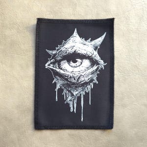 Op de afbeelding: Zwart-wit geborduurde patch met een gedetailleerd beeld van een groot, druppelend oog met een gekartelde, stekelige omtrek.