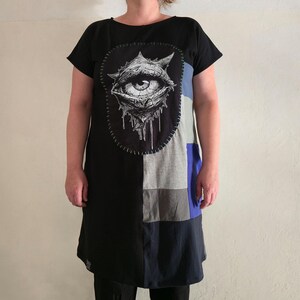 Può includere: Abito tunica nero e color block con un design grafico a occhio. L'abito a maniche corte presenta una grande grafica dettagliata dell'occhio sul davanti, con un design patchwork di pannelli neri, grigi e blu. La grafica dell'occhio è in bianco e nero.
