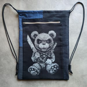 Pode incluir: Mochila com cordão preta e jeans. A frente apresenta um urso ameaçador segurando uma faca. Inclui um bolso com zíper e alças pretas.