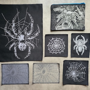 Pode incluir: Uma coleção de patches de aranha em preto e branco e uma bolsa com zíper. Os patches apresentam desenhos de teias de aranha e ilustrações detalhadas de aranhas. A bolsa tem um gráfico de tarântula e o texto "GOES FREE".