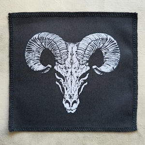Könnte beinhalten: Ein schwarzer Stoff-Patch mit einem weißen Bild eines Widder-Schädels mit großen Hörnern.