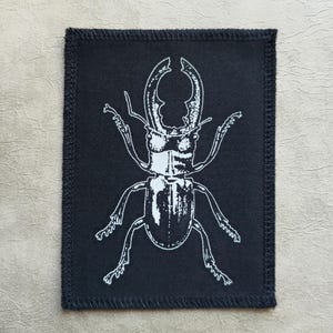 Op de afbeelding: Zwarte stoffen patch met een witte omtrek van een hertenkever. De kever kijkt naar voren met zijn poten uitgestrekt.