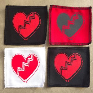 Puede incluir: Cuatro parches para planchar con un diseño de corazón roto. Los parches son de diferentes colores: negro, blanco, rojo y gris. El corazón es rojo en los parches negro y blanco, y gris en el parche rojo.