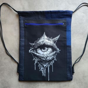 Op de afbeelding: Zwarte rugzak met trekkoord en een zwart-witte oogafbeelding. De rugzak heeft een blauw ritsvak en zwarte trekkoorden. De tas is gemaakt van denim stof.