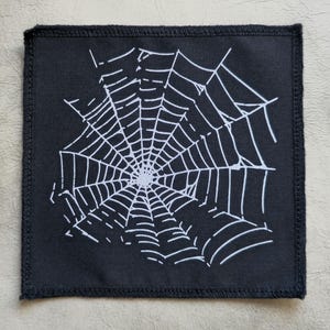 Peut inclure: Patch en tissu noir avec un motif de toile d'araignée blanche. La toile est détaillée et complexe, avec un point central et des lignes rayonnantes.