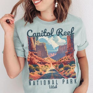May include: A light blue t-shirt featuring a scenic illustration of Capitol Reef National Park in Utah. The design includes a road winding through a canyon with red rock formations, blue skies, and the text "Capitol Reef National Park Utah."