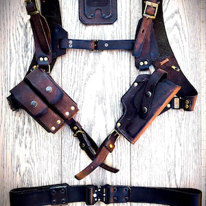 May include: A brown leather chest rig with two magazine pouches, a holster, and a belt. The rig has brass hardware and is designed to be worn over a shirt.