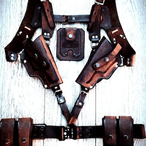 Pode incluir: Um coldre de arma de couro marrom com dois coldres para pistolas, um coldre para carregador e um cinto com duas bolsas adicionais. O coldre tem correias e fivelas ajustáveis.