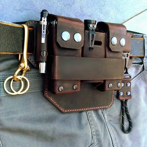 May include: A brown leather belt organiser with multiple pouches and compartments. The organiser features silver rivets, a pen holder, and a flashlight clip. A gold-coloured hook and key rings are attached to the belt.