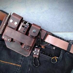 Puede incluir: Un cinturón de herramientas de cuero marrón con múltiples bolsillos y compartimentos, sujeto a un cinturón de cuero marrón. El cinturón se lleva sobre unos vaqueros oscuros. Los bolsillos contienen varias herramientas y accesorios.