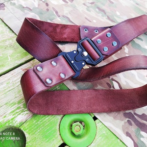 Könnte beinhalten: Ein brauner Ledergürtel mit einer schwarzen Metallschnalle und silbernen Nieten. Der Gürtel ist in der Mitte gefaltet und liegt auf einer grün-braunen Oberfläche.