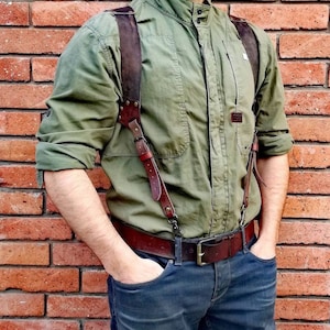 May include: A person wearing a green collared shirt with brown leather braces and a brown leather belt. The person is wearing blue jeans and has their hands in their pockets.