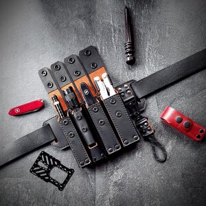 May include: A black leather belt with multiple pouches holding various tools and accessories. The pouches are secured with black snaps and feature a contrasting tan interior. A red Swiss Army knife and a red leather accessory are also visible.