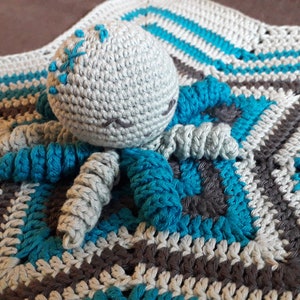 Può includere: Un giocattolo di polpo all'uncinetto con filato grigio e blu, sdraiato su una coperta all'uncinetto grigia, blu e marrone.