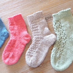 Può includere: Quattro calzini lavorati a mano a maglia nei toni del blu, rosa, grigio e verde. Ogni calzino presenta un motivo unico, tra cui punti strutturati e disegni traforati. I calzini sono esposti su una superficie di legno.