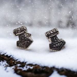 Pode incluir: Dois brincos prateados em forma de martelo de Thor, cada um com detalhes intrincados, repousam sobre uma superfície nevada. Os brincos têm a forma de Mjolnir, o martelo mítico. O fundo está desfocado, sugerindo uma cena de inverno.