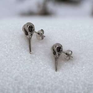 Op de afbeelding: Een paar zilveren schedel oorbel studs met zwarte oogdetails. De oorbellen liggen op een wit oppervlak.