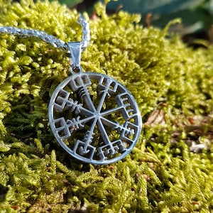 Puede incluir: Un collar con un colgante de plata en forma de br&uacute;jula circular adornado con un s&iacute;mbolo Vegvisir y runas. El collar est&aacute; sobre un lecho de musgo verde.