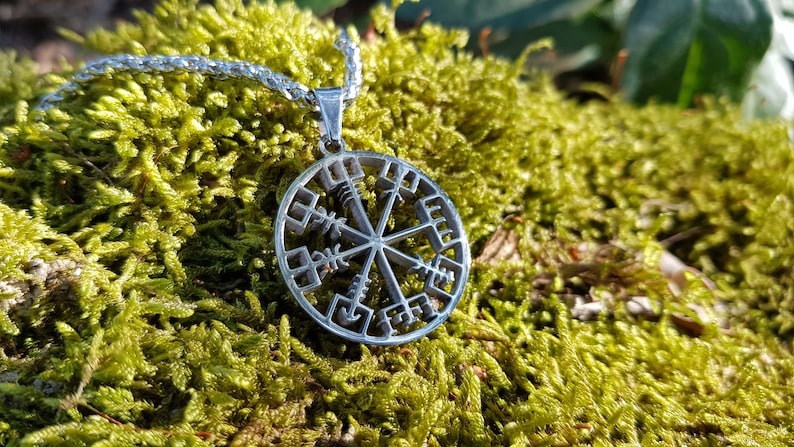 Puede incluir: Un collar con un colgante de plata en forma de br&uacute;jula circular adornado con un s&iacute;mbolo Vegvisir y runas. El collar est&aacute; sobre un lecho de musgo verde.