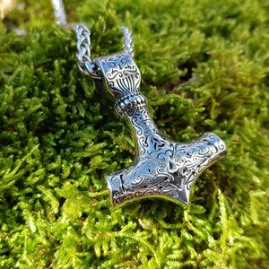 Op de afbeelding: Een zilveren Mjolnir-hanger met ingewikkelde bloemmotieven, hangend aan een zilveren ketting. De hanger ligt op een bed van groen mos.