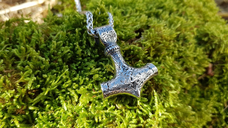 Op de afbeelding: Een zilveren Mjolnir-hanger met ingewikkelde bloemmotieven, hangend aan een zilveren ketting. De hanger ligt op een bed van groen mos.