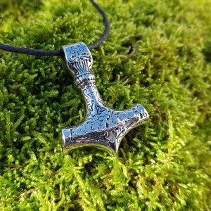 Op de afbeelding: Een zilveren Mjolnir-hanger met ingewikkelde bloemmotieven, hangend aan een zwart koord tegen een groene mossige achtergrond.