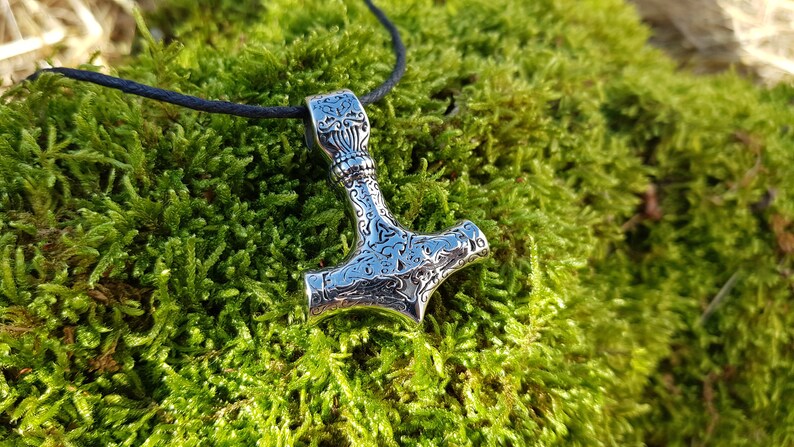Op de afbeelding: Een zilveren Mjolnir-hanger met ingewikkelde bloemmotieven, hangend aan een zwart koord tegen een groene mossige achtergrond.