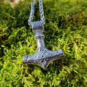 Op de afbeelding: Een zilveren Mjolnir-hangerketting met ingewikkelde gegraveerde details. De hanger hangt aan een zilveren ketting tegen een groene mossige achtergrond.