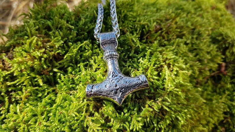 Op de afbeelding: Een zilveren Mjolnir-hangerketting met ingewikkelde gegraveerde details. De hanger hangt aan een zilveren ketting tegen een groene mossige achtergrond.