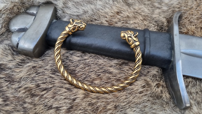 May include: A gold-toned Viking-style bracelet with two wolf head terminals. The bracelet is made of twisted metal and has a braided design. The bracelet is lying on a brown fur surface.